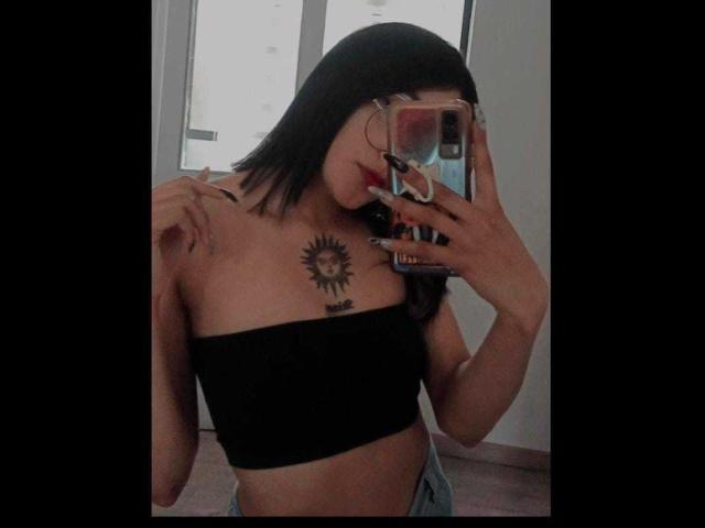 SalomeMille with black hair, red lipstick, black tube top, sun tattoo on chest, holding phone, mirror selfie, visible ass, medium breasts, slight tan, casual room.
