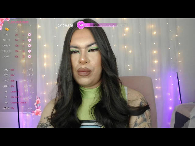 ImNicol has long black hair, green eyeshadow, and a green turtleneck. ImNicol's tattoos are visible on her arms, and she has a pouty expression. Her ass is not visible.