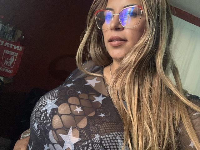 TompsonKarina with long blonde hair, wearing reflective glasses and a sheer black top with white star patterns, exposing her big tits. She's smiling slightly.