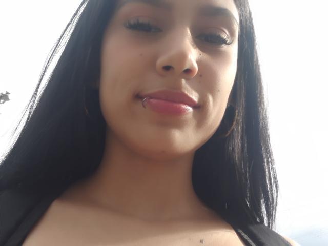 KataleyaMansson has long black hair, brown eyes, and a lip piercing. She's wearing hoop earrings and has glossy pink lips, looking down at the camera with a slight smile.