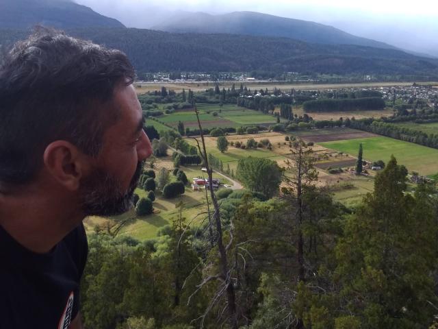 Jpaolo69 with a beard, side profile, looking down at a green, hilly landscape with a small town and fields. He's wearing a black shirt, and it's a daytime shot.