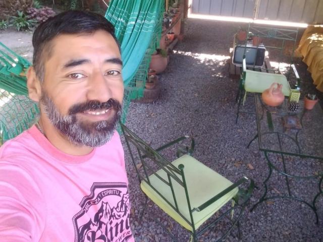 Jpaolo69 has a medium build, short black hair, and a gray beard, wearing a pink shirt, smiling, with green metal chairs and potted plants in a gravel yard.