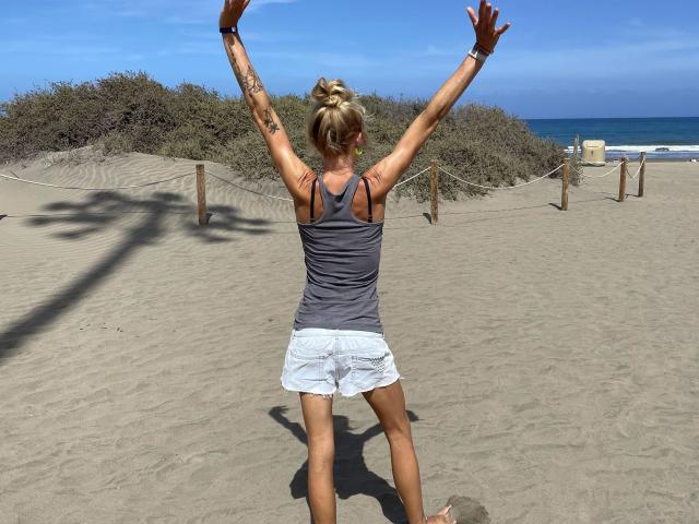 Rojia-hot stands on a beach with arms raised, wearing a gray tank top and white shorts, showing tattoos on both arms, and has blonde hair in a bun.