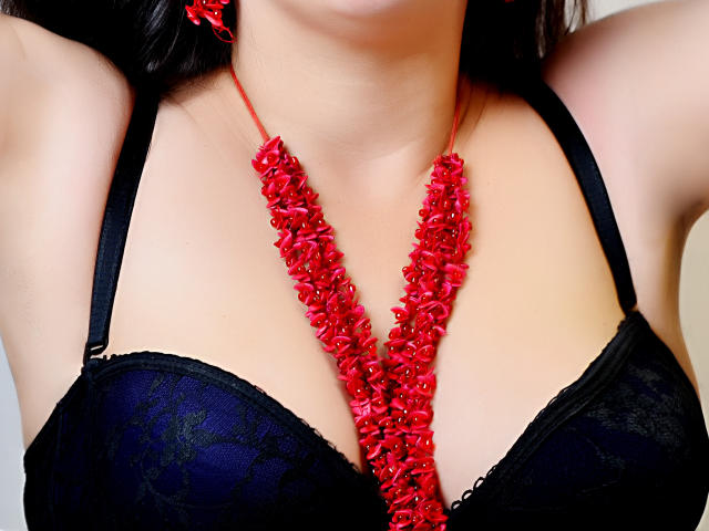 PriscillaHotMature's black lace bra, with red rose necklace, highlighting her tits and pale skin. She's wearing red rose earrings, arms raised, showing her cleavage.