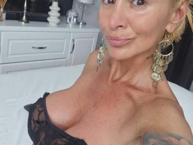 BriliantOne with large, exposed tits in black lace, wearing big, ornate earrings, smiling at the camera, showing a tattoo on her right arm. Blonde, slightly tanned, bedroom setting.