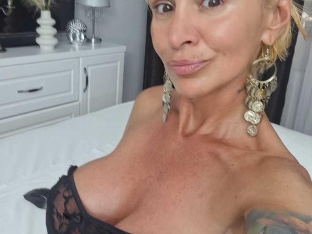 BriliantOne with blonde hair, wearing big gold earrings and black lace bra, showing off her tits, smiling at the camera, lying on a white bed.