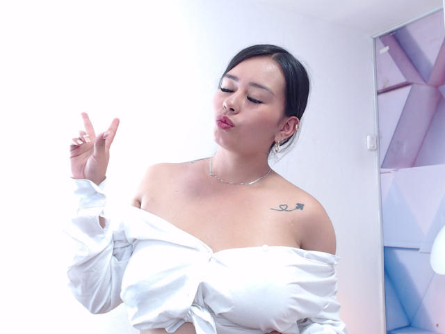 MelissaCaty com lábios curvados, cabelo preto, blusa branca com um ombro descoberto, seios pequenos, tatuagem no ombro direito, dedo levantado, colar de prata e brincos, em um quarto bem iluminado..