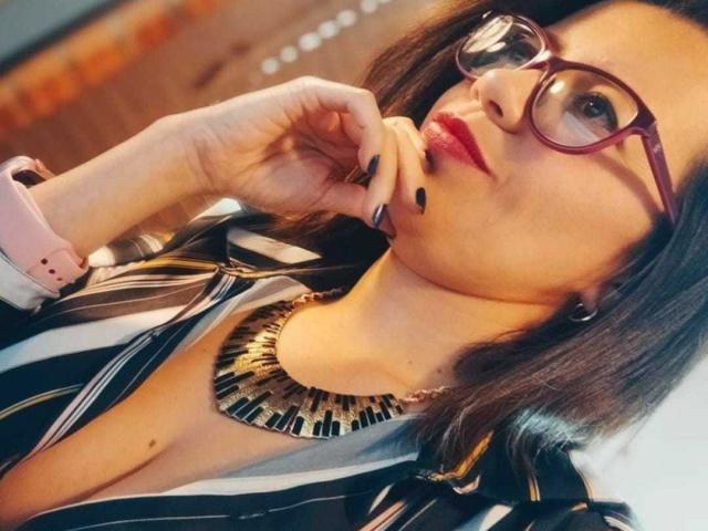 AbbyCollings met rode lippenstift, een bril en zwarte nagellak, draagt een gestreepte shirt, een roze polsbandje en een dikke ketting, en raakt aan haar kin. Haar borsten zijn zichtbaar..