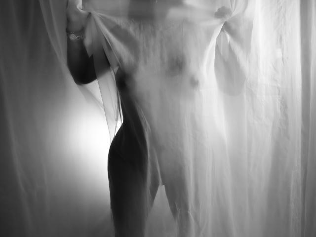 LizHerrera's naked body is partially covered by a sheer, white curtain. Her small tits and slim, toned ass are visible. Her hand holds the curtain. Black-and-white photo.
