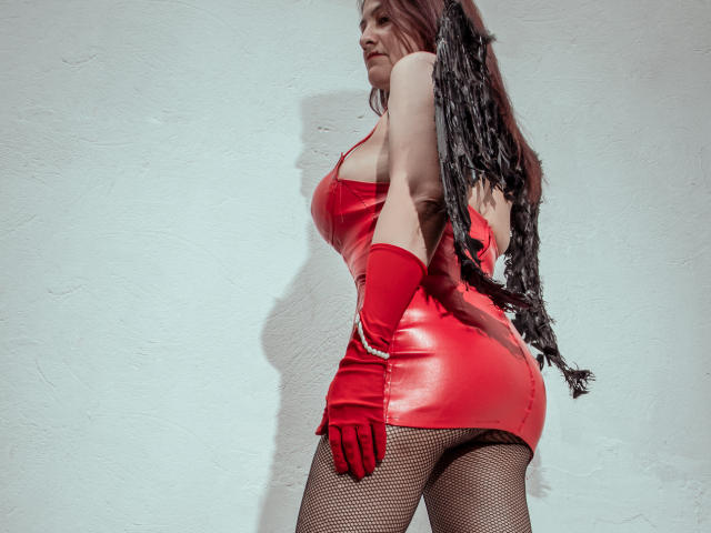 MilenneSexy in a tight red latex dress, red gloves, fishnet stockings, and long black braids, showing off her ass and curvy figure, with a confident smile.