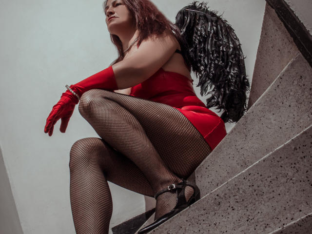 MilenneSexy squats on stone steps, wearing red lingerie, fishnet stockings, red gloves, black heels, and black feather wings. Her ass is prominent, and she has red hair.