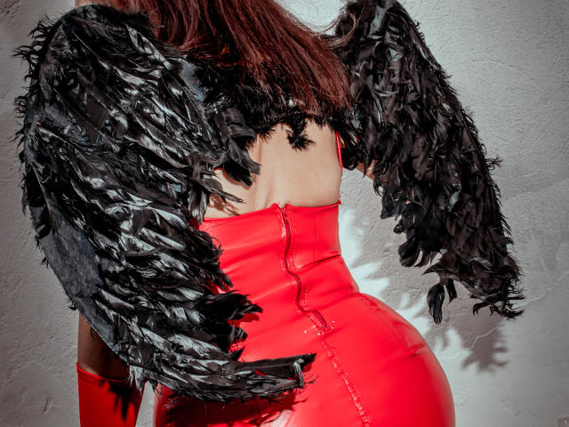 MilenneSexy in a shiny red latex dress with black feathered wings, back facing camera, showing her ass and upper back. She has brown hair, standing against a white wall.