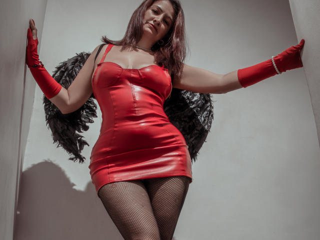 MilenneSexy in a red leather dress, black fishnet stockings, red gloves, and black feather wings, standing with arms out, looking down at the camera, showing her tits.