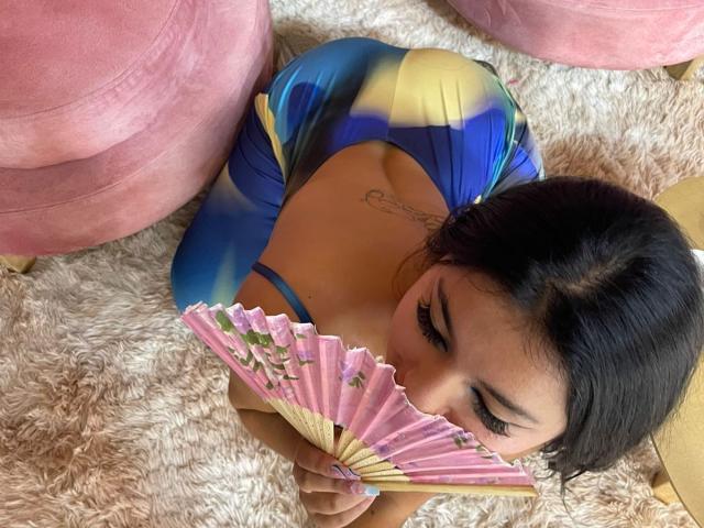 AlessiaFerry, met donker haar, een blauw-gele jurk en een zichtbare tatoeage, knielend op een pluizig tapijt, met een roze waaier voor haar borsten, en omhoog kijkend met lange wimpers..