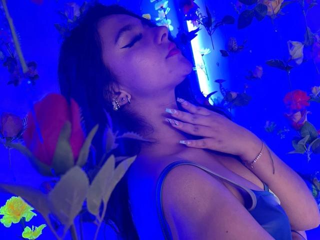 AlessiaFerry with blue-lit skin, eyes closed, hand on neck, wearing a blue tank top, floral earrings, and a bracelet, surrounded by flowers, looking sexy and relaxed.
