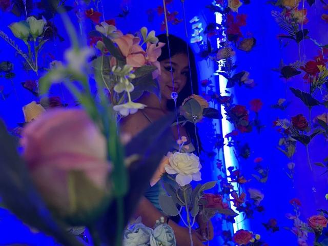 Le visage d'AlessiaFerry est partiellement dissimulé par des fleurs colorées, avec des roses roses et blanches au premier plan. Un éclairage bleu met en valeur ses cheveux foncés et son sourire discret..