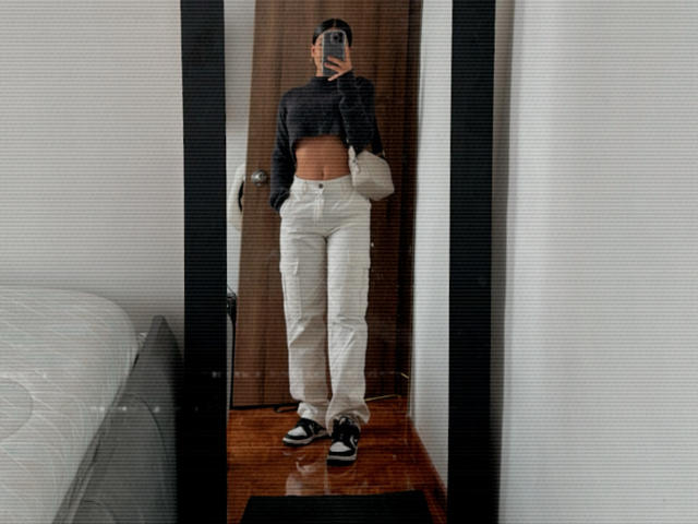 ThaliaAres with a flat stomach, wearing a black crop top, white pants, black and white sneakers, and a beige bag, taking a mirror selfie.