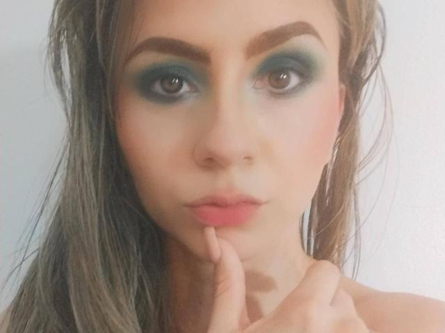 KariCarter has teal eye makeup, brown eyebrows, pink lips, and touches her chin with her finger. She has wet, brown hair and a neutral expression.