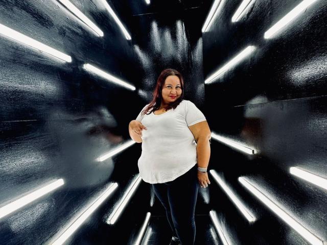 AliceShae stands in a black, shiny hallway with white lights, wearing a white shirt, black pants, and a watch, with a slightly chubby, curvy body and a red smile.