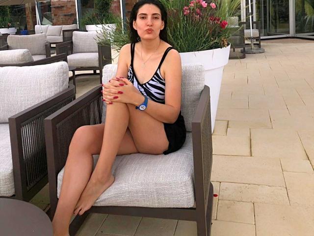 AddictedToUTS with zebra-patterned top, black shorts, and red nails, sitting on a gray cushioned chair, pouting, legs crossed, wearing a blue wristwatch.