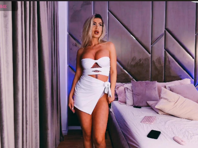 Lellie stands in a bedroom wearing a white strapless crop top and skirt, exposing her tits and a cutout on her midriff. Her ass is visible. Bed with pillows.