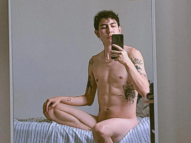 Naked BlakeTyler with tattoos, sitting cross-legged on a bed, taking a mirror selfie with a phone. BlakeTyler has a slim, muscular build and short curly dark hair.