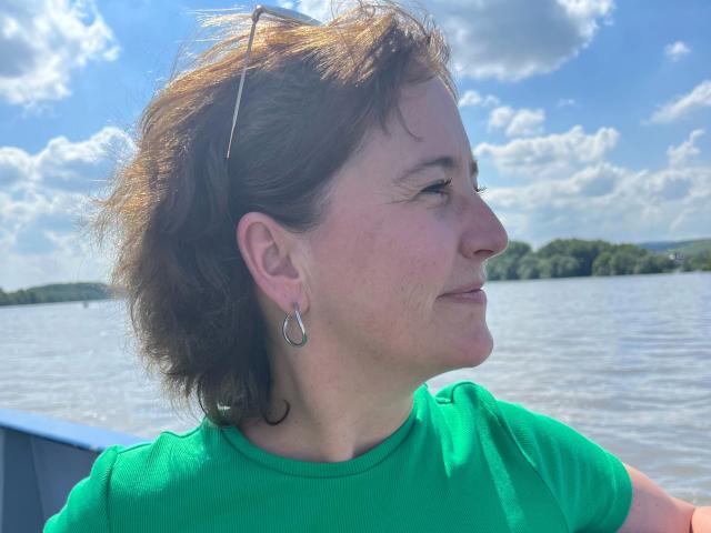 Pamm2023-hot with short brown hair, green shirt, hoop earrings, and sunglasses on head, looking side profile with a slight smile, standing on a boat with a river behind.