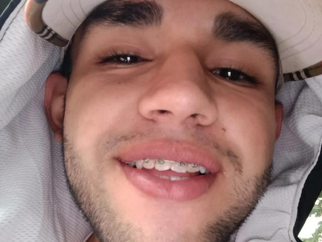TonnyMonthana's face with short dark hair, brown eyes, light skin, and a slight beard, smiling with metal braces on his teeth, wearing a white hat and towel.