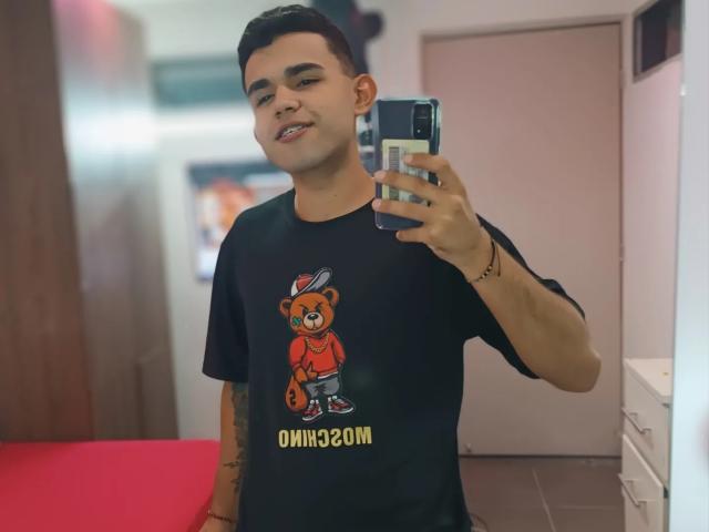 TonnyMonthana, met kort zwart haar, draagt een zwart T-shirt met een schattige afbeelding van een teddybeer, houdt een telefoon vast in een spiegelzelfportret, en glimlacht terwijl hij tatoeages op zijn arm heeft..