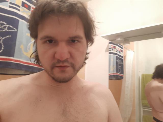 NectarineFad, sem camisa, com cabelo castanho despenteado e barba, tem o peito peludo e está olhando para a câmera. Seu reflexo é visível no espelho..