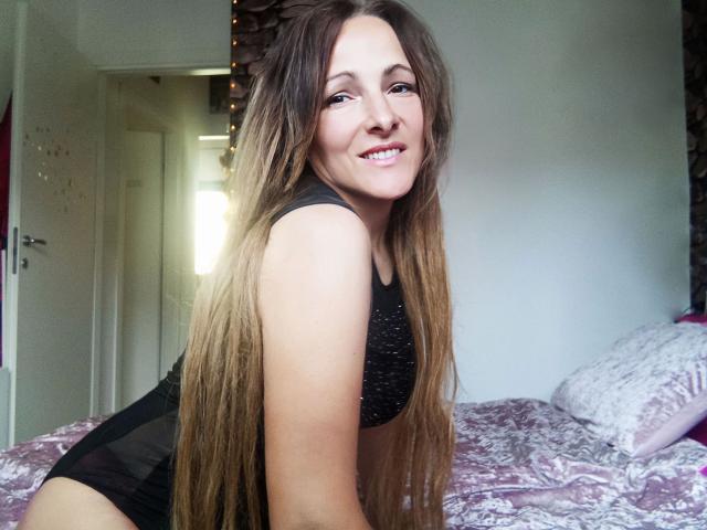 Una Vivien matura, attraente, con lunghi capelli castani, sorridente, che indossa un reggiseno sportivo nero e biancheria intima nera, inginocchiata su un letto con lenzuola a motivi rosa e bianchi. Il seno è visibile..