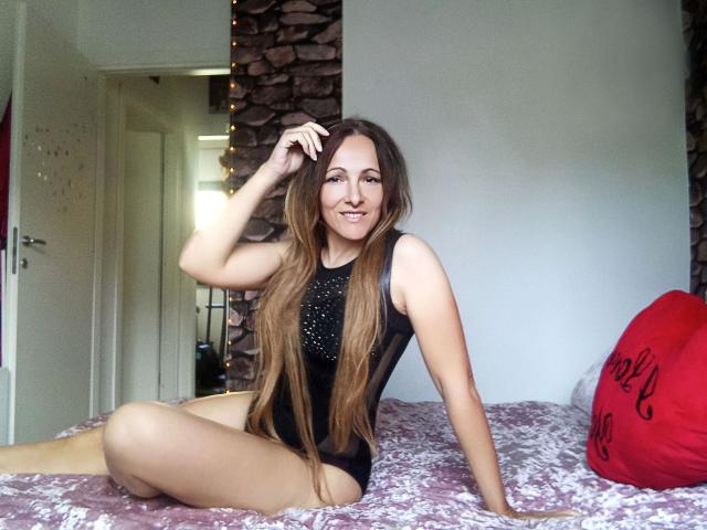 ReifeVivien-hot with long brown hair, smiling, wearing a black one-piece with studs, sitting on a bed, hand in hair, legs crossed, pink and white bedspread, red heart pillow.