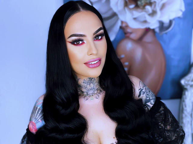 HottyNicole has long black hair, red eye makeup, tattoos on her neck and arm, and a black lace top. She's smiling with pink lipstick and showing her tits.