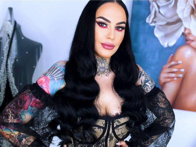 HottyNicole with long black hair, red eyeshadow, and tattoos, wearing a black lace top, showing off her tits and colorful arm tattoos, with a confident smile.