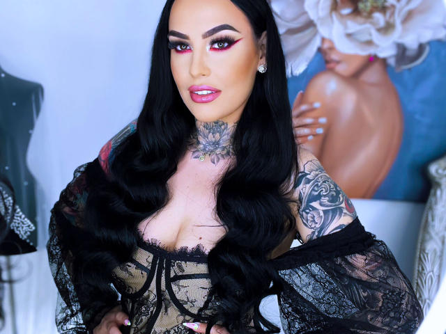 HottyNicole with black lace dress, showing tits, tattoos on neck and arm, long black hair, pink eye makeup, and pink lipstick, standing with hands on hips.