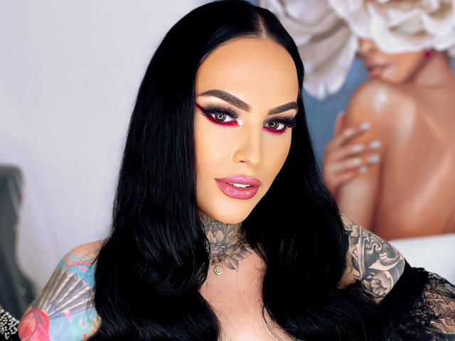 HottyNicole has long black hair, red eyeshadow, and glossy pink lips. She's tattooed with colorful designs on her arms and has large, shiny tits. She's wearing lace.