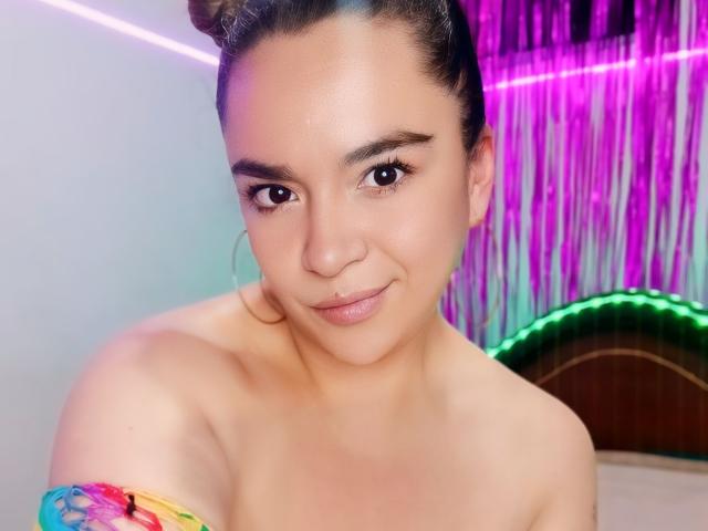 NaugthyCurvy with light brown skin, dark hair in buns, small nose, subtle smile, hoop earrings, colorful off-shoulder top, showing bare shoulders and upper chest, pink and purple lights.