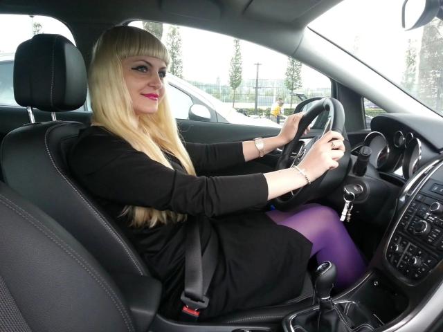 MistresDesiree-hot with blonde hair, black dress, purple tights, and red lipstick, sits in a car, gripping the steering wheel, wearing a seatbelt, with a sexy smile.