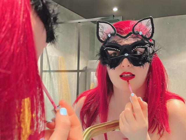 MelanieFrancaiseDuo mit hellroten Haaren, schwarzer Katzenmaske mit Ohren und rotem Lippenstift, putzt mit einer rosa Zahnbürste ihre Zähne, wobei sich das Spiegelbild darin zeigt, und hält einen goldenen Spiegelgriff..
