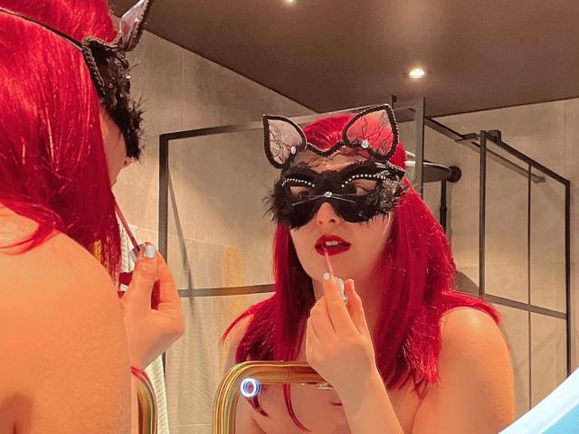 MelanieFrancaiseDuo: Eine Frau mit roten Haaren, einer schwarzen Katzenmaske und rotem Lippenstift, die mit dem Finger auf ihre roten Lippen tippt, wobei sie sich in einem Badezimmerspiegel leicht entblößt..