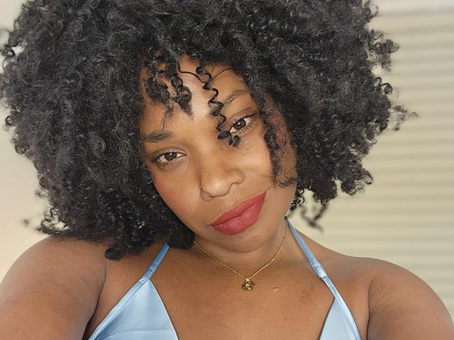 RachelDiamond has dark curly hair, brown eyes, red lipstick, and a light blue halter top. She's wearing a gold necklace with a small pendant. Her tits are small.