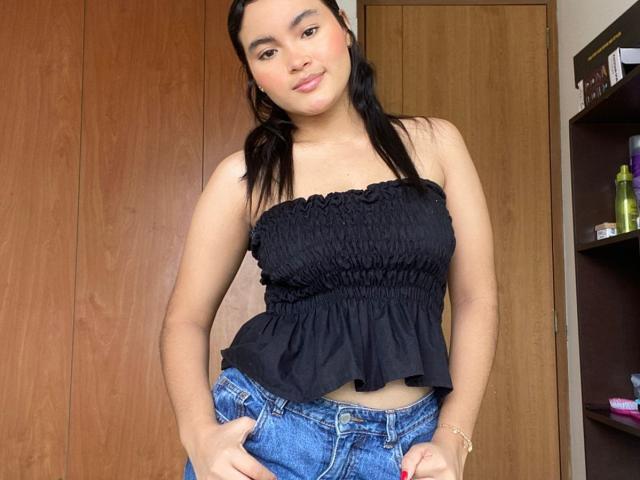 MiiaPalmer with dark hair, wearing a black strapless top, exposing small tits, and blue jeans, standing in front of wooden doors, smiling with hands on her hips.