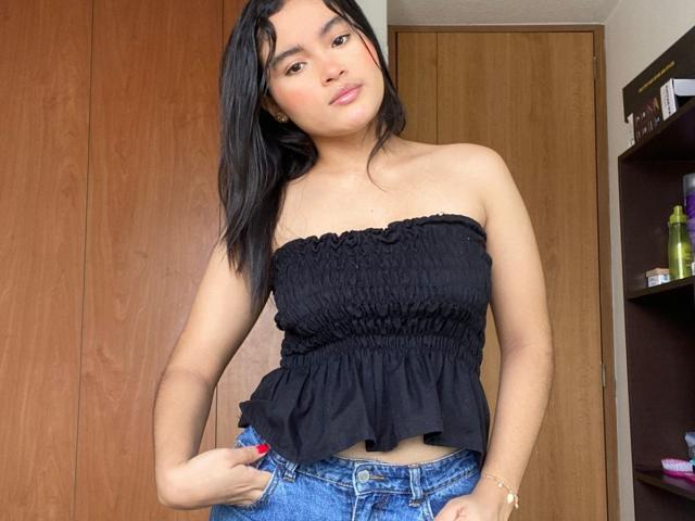 MiiaPalmer with black strapless top, ruffled midsection, and blue jeans, standing, hand on hip, smiling, long black hair, light brown skin, small tits, red nail polish, wooden door.