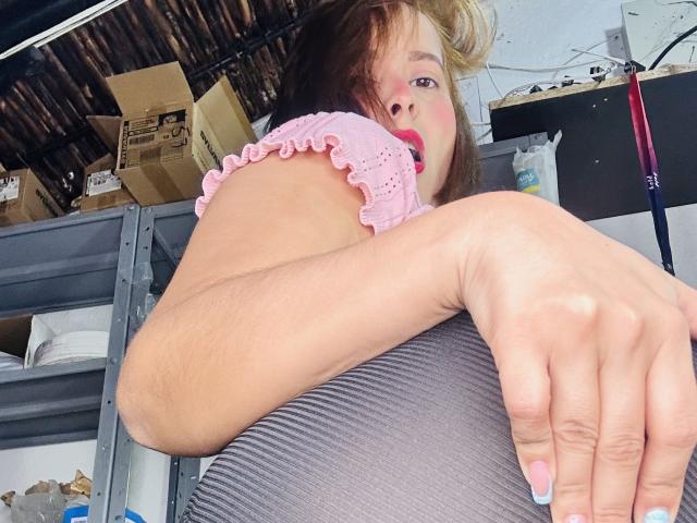 SaritaVelez hugs a gray pillow with her right arm, wearing a pink ruffled top, her blonde hair partially covering her face, showing red lipstick and a slight smile.