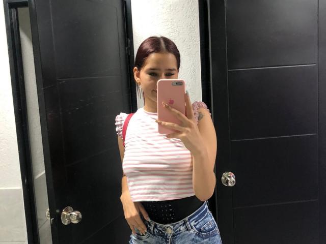 MelodyMansson taking a mirror selfie, wearing a pink-striped top, black belt, and blue jeans, with her hand on her ass, holding a pink phone, in a black room.