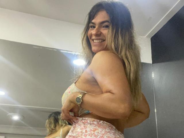 BrianaPreston with long, wavy brown hair, smiling, wearing a floral bra, showing her toned, muscular arms, and pink floral pants, wearing a watch, in a mirror selfie.