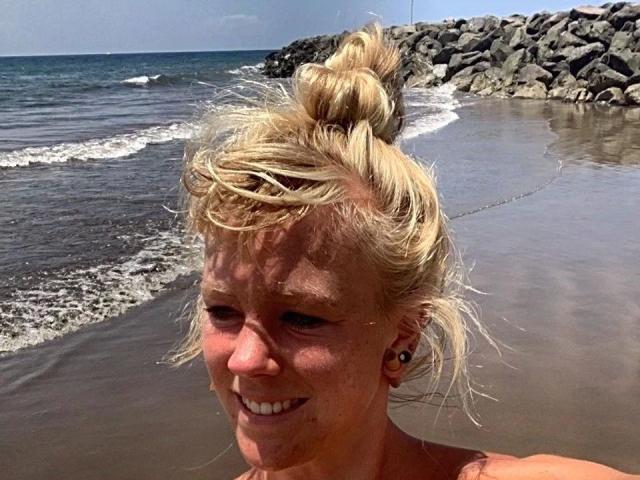 Rojia-hot has blonde hair in a messy bun, sunburned skin, and a small smile, standing at a rocky beach with waves in the background.