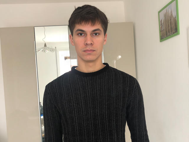 VioletaAndLeo with short brown hair, wearing a black, textured, long-sleeve sweater, standing in a room with a mirror and light walls, looking straight at the camera.