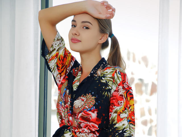 VioletaAndLeo with a hand on her head, wearing a colorful floral robe, smiling, brown hair in a ponytail, light skin, and pink lipstick, standing by a window.