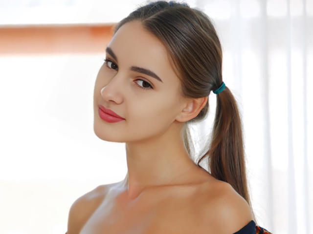 VioletaAndLeo has a cute face with brown eyes, pink lips, and a small nose, wearing a blue hair tie in a brown ponytail, showing bare shoulders.